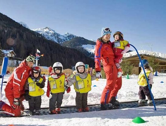 Zwei Erwachsene und fünf Kinder in Skiausrüstung stehen im Schnee; die Kinder tragen gelbe Westen und Helme, im Hintergrund sind Berge und Bäume zu sehen.