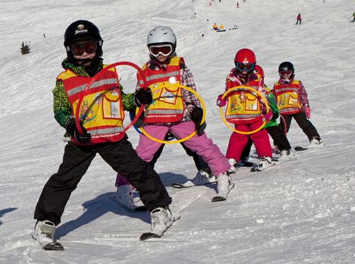 Fünf Kinder mit Helmen und Sicherheitswesten fahren in einer Reihe auf einer verschneiten Piste Ski, jedes hält einen farbigen Ring.