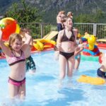 Kinder und Erwachsene spielen mit aufblasbarem Spielzeug in einem kleinen Freibad an einem sonnigen Tag, mit Bergen im Hintergrund.