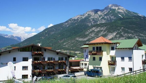 Eine Gruppe von Gebäuden im alpinen Stil mit Blumenkästen liegt am Fuße eines grünen Berges unter einem klaren blauen Himmel.