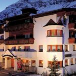 Ein mehrstöckiges alpines Hotel mit Holzbalkonen und weißen Wänden vor dem Hintergrund schneebedeckter Berge wird in der Abenddämmerung mit einer Weihnachtsmannfigur am Eingang beleuchtet.