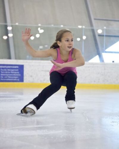 Ein junges Mädchen in einem rosafarbenen Tank-Top und einer schwarzen Hose führt eine niedrige Eislaufbewegung auf einer überdachten Eislaufbahn aus.
