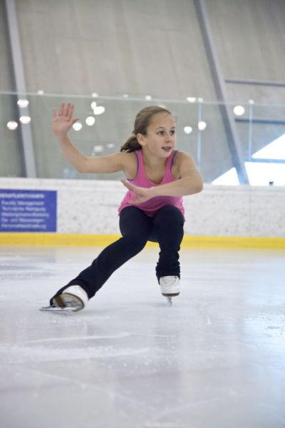 Ein junges Mädchen in einem rosafarbenen Tank-Top und einer schwarzen Hose führt eine niedrige Eislaufbewegung auf einer überdachten Eislaufbahn aus.