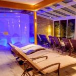 Moderner Entspannungsbereich im Innenbereich mit Lounge-Sesseln aus Korbgeflecht, einer Glaswand, Pflanzen, Steinakzenten und blauer Stimmungsbeleuchtung, die eine spaähnliche Umgebung schafft.