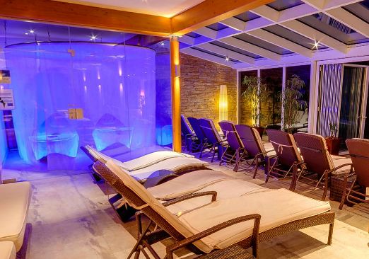 Moderner Entspannungsbereich im Innenbereich mit Lounge-Sesseln aus Korbgeflecht, einer Glaswand, Pflanzen, Steinakzenten und blauer Stimmungsbeleuchtung, die eine spaähnliche Umgebung schafft.