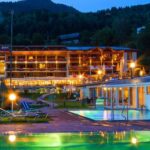 Ein hell erleuchtetes Hotel mit Holzbalkonen ist in der Abenddämmerung zu sehen, umgeben von Bäumen und Hügeln, mit Außenpools und Liegestühlen im Vordergund.