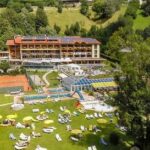 Ein großer Hotelkomplex mit Außenpools, Tennisplätzen und Liegestühlen ist von grünen Rasenflächen und Bäumen in einer hügeligen Gegend umgeben.