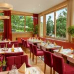 Ein heller Restaurantbereich mit roten Stühlen, gedeckten Tischen mit gefalteten Servietten, großen Fenstern und Topfpflanzen.