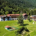 Eine Ansammlung von Gebäuden liegt auf einem grünen Hügel, umgeben von dichtem Wald und offenen Grasflächen, mit einem eingezäunten Bereich und kleinen Gebäuden in der Nähe.