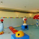 Indoor-Kinderbecken mit flachem Wasser und bunten Spielstrukturen, darunter eine Robbe mit Ball, ein Clown, ein Pilzbrunnen und eine gelbe Figur.