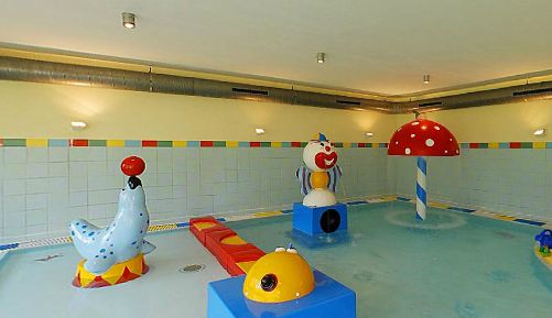 Indoor-Kinderbecken mit flachem Wasser und bunten Spielstrukturen, darunter eine Robbe mit Ball, ein Clown, ein Pilzbrunnen und eine gelbe Figur.