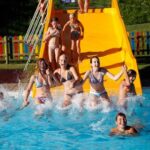 Eine Gruppe von Kindern in Badeanzügen spritzt von einer gelben Rutsche in ein Becken, während andere oben warten. Ein bunter Zaun umgibt den Bereich.