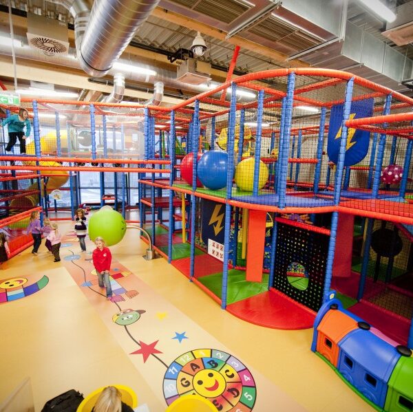 Kinder spielen in einem großen Indoor-Spielplatz mit bunten Klettergerüsten, Rutschen, Netzen und weichen Spielgeräten bei heller Beleuchtung.