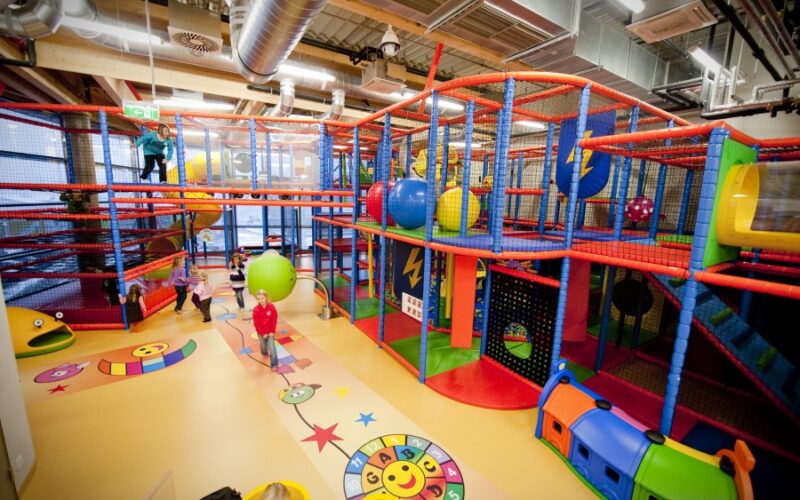 Kinder spielen in einem großen Indoor-Spielplatz mit bunten Klettergerüsten, Rutschen, Netzen und weichen Spielgeräten bei heller Beleuchtung.