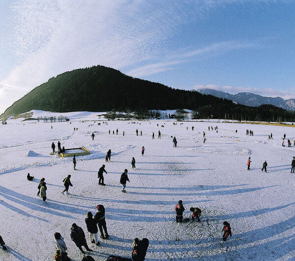 Eine weite Ansicht von Menschen, die auf einem großen zugefrorenen See Schlittschuh laufen und spazieren gehen, mit einem bewaldeten Hügel und Bergen im Hintergrund unter einem klaren Himmel.