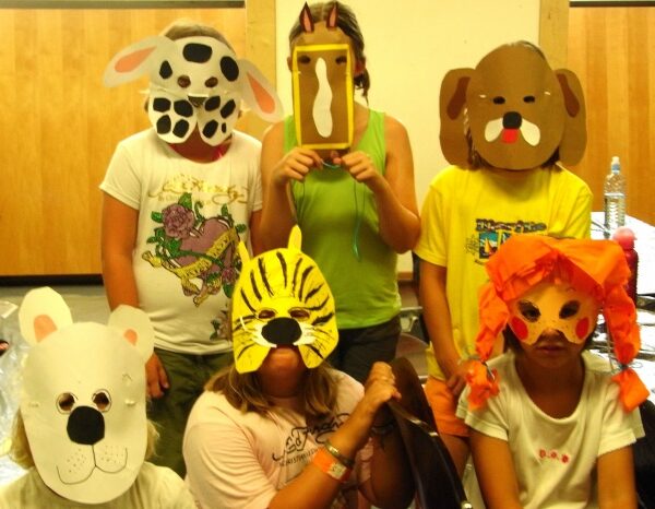 Sechs Kinder tragen handgefertigte Tiermasken und sitzen bzw. stehen gemeinsam im Raum und posieren für das Foto. Im Hintergrund sind Holzvertäfelungen und ein Tisch zu sehen.