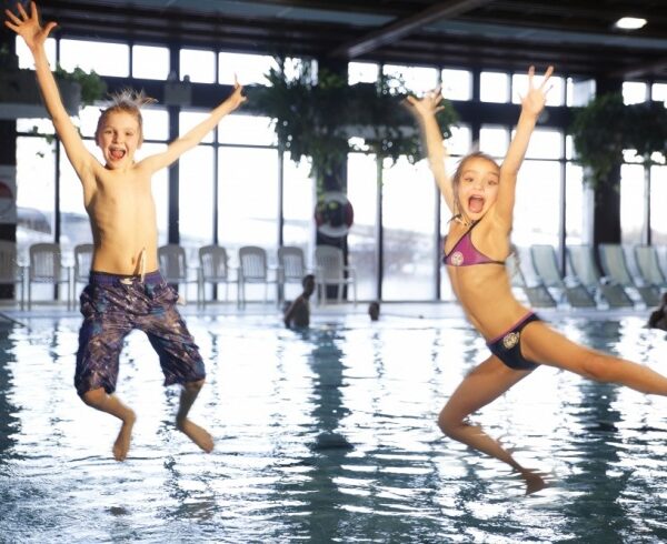 Zwei Kinder in Badeanzügen springen in ein Hallenbad. Um sie herum plätschert das Wasser. Im Hintergrund sind weitere Personen zu sehen.