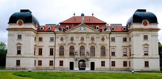Großer Palast im Barockstil mit verzierter Fassade, bogenförmigem Eingang, rotem Dach, zwei Kuppeltürmen und symmetrischen Fenstern, auf einer Wiese unter einem bewölkten Himmel.