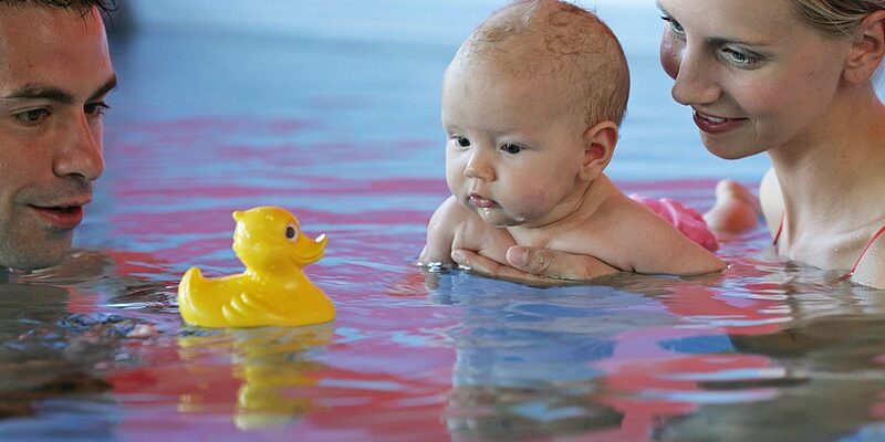 Zwei Erwachsene halten ein Baby in einem Schwimmbecken, während das Baby eine gelbe Gummiente ansieht, die auf dem Wasser schwimmt.