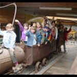 Eine Gruppe von Kindern sitzt und steht auf einem alten Bergwerkswagen in einem Holzgebäude, winkt und lächelt in die Kamera.