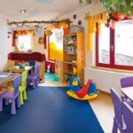 Buntes Kinderspielzimmer mit kleinen Tischen und Stühlen, Spielzeug, Bücherregalen, Spielgeräten und hellem Dekor; natürliches Licht fällt durch die Fenster.