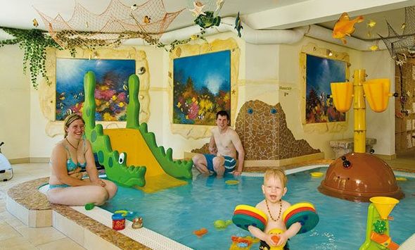 Drei Personen, darunter ein Kind in einem Schwimmer, spielen in einem Kinderbecken mit Wasserspielzeug und einer grünen Rutsche, umgeben von farbenfroher Wasserdekoration.