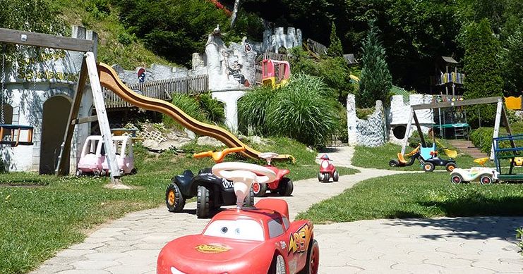 Spielplatz im Freien mit Spielzeugfahrzeugen auf einem Weg, Spielgeräten im Stil einer Burg, Rutschen und Grünflächen im Hintergrund.