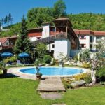 Ein kleiner Außenpool mit Liegestühlen, Sonnenschirmen, einer Spielplatzrutsche und Grünflächen neben einem Hotelgebäude in einer hügeligen Gegend.