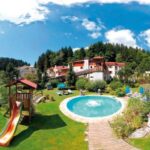 Rundes Freibad und Spielplatz mit Rutsche auf einer Wiese, umgeben von Liegestühlen, Bäumen und Gebäuden unter einem blauen Himmel mit Wolken.