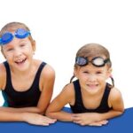 Zwei kleine Kinder mit Schwimmbrillen und schwarzen Badeanzügen lehnen auf einer blauen Fläche und lächeln in die Kamera vor einem weißen Hintergrund.