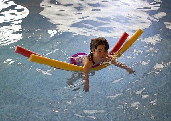 Ein junges Mädchen schwimmt in einem Hallenbad mit zwei Schaumstoffnudeln als Stütze.
