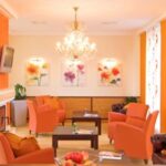 Helle Lounge mit orangefarbenen Stühlen, floraler Wandkunst, einem Kronleuchter und gemusterten Vorhängen. Auch Couchtische und ein Obstkorb sind zu sehen.