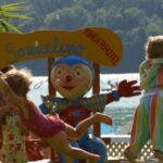 Erwachsene und Kinder spielen in der Nähe eines farbenfrohen Clownsschildes an einem Seeufer, während im Hintergrund Seifenblasen in der Luft schweben und Grünpflanzen zu sehen sind.