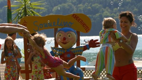 Erwachsene und Kinder spielen in der Nähe eines farbenfrohen Clownsschildes an einem Seeufer, während im Hintergrund Seifenblasen in der Luft schweben und Grünpflanzen zu sehen sind.