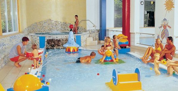 Kinder und Erwachsene spielen und entspannen sich in einem flachen Innenbecken mit buntem Spielzeug und einem kleinen Whirlpool im Hintergrund.