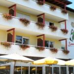 Ein dreistöckiges Hotel mit Blumenkästen auf jedem Balkon, einer Sonnenkarikatur und einem Logo an der Wand sowie gelben Terrassenschirmen im Vordergrund.