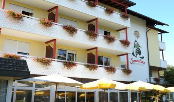 Ein dreistöckiges Hotel mit Blumenkästen auf jedem Balkon, einer Sonnenkarikatur und einem Logo an der Wand sowie gelben Terrassenschirmen im Vordergrund.