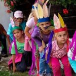 Eine Gruppe von Kindern mit bunten Federstirnbändern hockt in einer Reihe im Freien und bereitet sich auf eine Aktivität vor. Ein Erwachsener sitzt hinter ihnen.