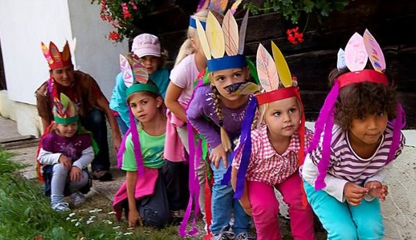 Eine Gruppe von Kindern mit bunten Federstirnbändern hockt in einer Reihe im Freien und bereitet sich auf eine Aktivität vor. Ein Erwachsener sitzt hinter ihnen.