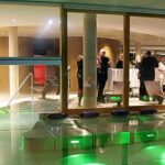 Eine Gruppe von Menschen steht und unterhält sich in einer modernen Lounge mit großen Glasfenstern, während grüne Lichter den Außenbereich in der Nähe eines Pools erhellen.