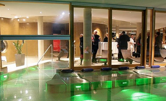 Eine Gruppe von Menschen steht und unterhält sich in einer modernen Lounge mit großen Glasfenstern, während grüne Lichter den Außenbereich in der Nähe eines Pools erhellen.