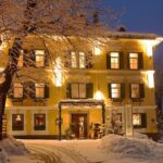 Ein gelbes, dreistöckiges Gebäude mit beleuchteten Fenstern steht an einem Winterabend inmitten schneebedeckter Bäume und Böden.