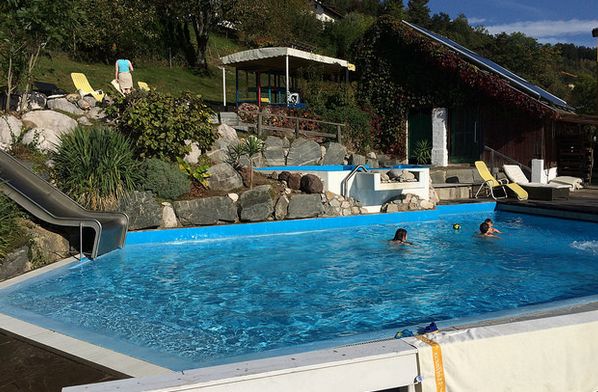 Zwei Personen schwimmen in einem rechteckigen Außenbecken mit einer Wasserrutsche, Felsen und Pflanzen in der Nähe. Im Hintergrund befinden sich Liegestühle und eine Struktur.