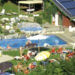 Ein kleiner Außenpool mit Liegestühlen, Sonnenschirmen, einer Rutsche, Sonnenkollektoren auf einem Gebäude und Menschen, die sich im und am Pool entspannen.