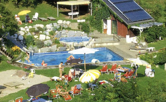 Ein kleiner Außenpool mit Liegestühlen, Sonnenschirmen, einer Rutsche, Sonnenkollektoren auf einem Gebäude und Menschen, die sich im und am Pool entspannen.