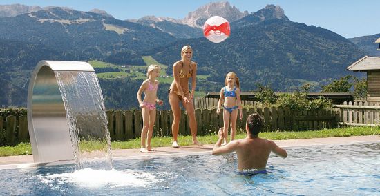 Eine Frau und zwei Mädchen spielen mit einem Strandball in einem Schwimmbad mit einem Mann, mit einer Berglandschaft im Hintergrund.