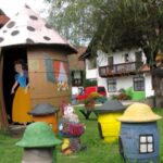 Eine kleine, mit Schneewittchen und einem Zwerg bemalte Hütte steht auf einer grasbewachsenen Wiese, umgeben von bunten Zwergenstatuen und nahe gelegenen Gebäuden.