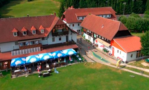 Luftaufnahme einer ländlichen Anlage mit roten Gebäuden, Sitzgelegenheiten im Freien unter Sonnenschirmen und einer von Bäumen umgebenen Rasenfläche.