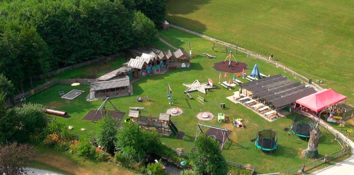 Luftaufnahme eines Spielplatzes im Freien mit verschiedenen Spielgeräten, Trampolinen, Schaukeln und kleinen Holzgebäuden auf einer Wiese in der Nähe eines Waldgebiets.