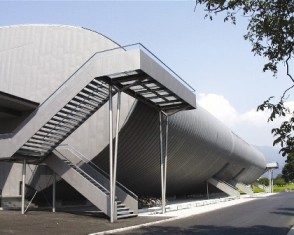 Außenansicht eines modernen Gebäudes im Industriestil mit einer großen zylindrischen Struktur und einer Außentreppe aus Metall, die zu einem erhöhten Gehweg führt.